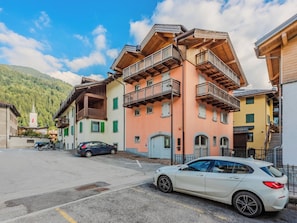 A multi-story building with balconies, a parking area, and a mountain backdrop. - Apartment Rugia by Interhome (Pinzolo)