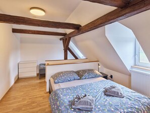 Ein Schlafzimmer mit schrägverdeckter Decke, Holzbalken, einem Bett mit Blumenbettwäsche, einem Nachttisch und einem Fenster.