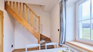 Una habitació amb una escala de fusta, una taula blanca amb un suport de tela teixida i una finestra amb vista a l'exterior.