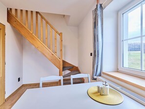 Ein Zimmer mit einer Holztreppe, einem weißen Tisch mit einer gewobenen Tischmatte und einem Fenster mit Blick ins Freie.