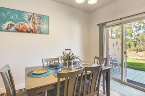 Un coin repas avec une table dressée pour quatre personnes, des chaises, un tableau accroché au mur et une vue à travers une porte vitrée.