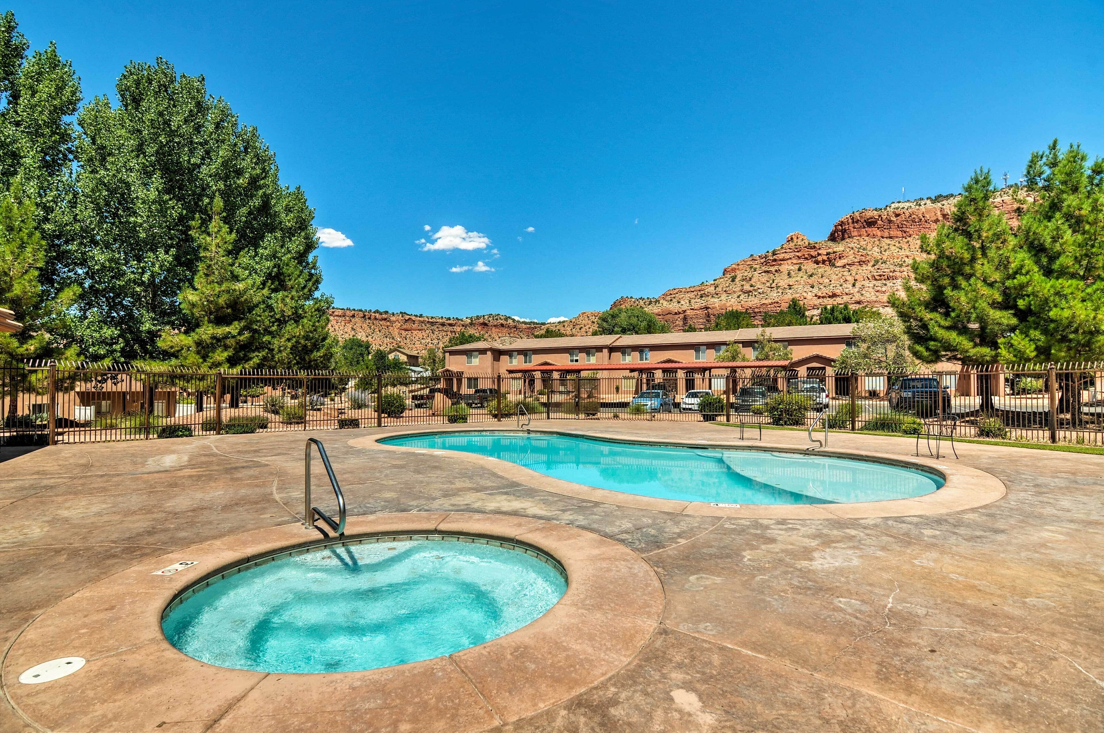 A pool area with multiple pools and a hot tub, surrounded by a fence and trees, with a building in the background.