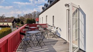 Um terraço ao ar livre com mesas e cadeiras, guarda-corpo vermelho e um edifício com fachada branca e porta de vidro.