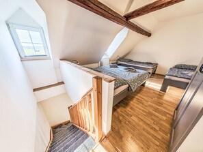 Un dormitorio tipo altillo, pequeño y acogedor, con techo inclinado, vigas de madera, un ventanal y una cama empotrada con colcha estampada.