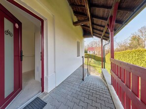 Ein überdachter Außenbereich mit roter Brüstung, einer Tür mit Fahrradsymbol und einem gepflasterten Weg, der zu einem Haus im Hintergrund führt.