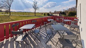 Een terras buiten met tafels en stoelen, een rode balustrade en uitzicht op het landschap.