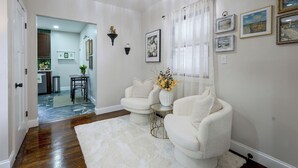 A room with a white rug, two white armchairs, a small table, and a white vase with flowers. There are framed pictures on the wall and a door leading to another room. - Walk to Water, Dining! Modern City Point Haven (New Haven)