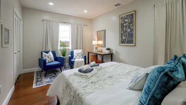 A hotel room with a bed, a seating area with chairs, a table with a lamp, a window with curtains, and a painting on the wall. - Walk to Water, Dining! Modern City Point Haven (New Haven)
