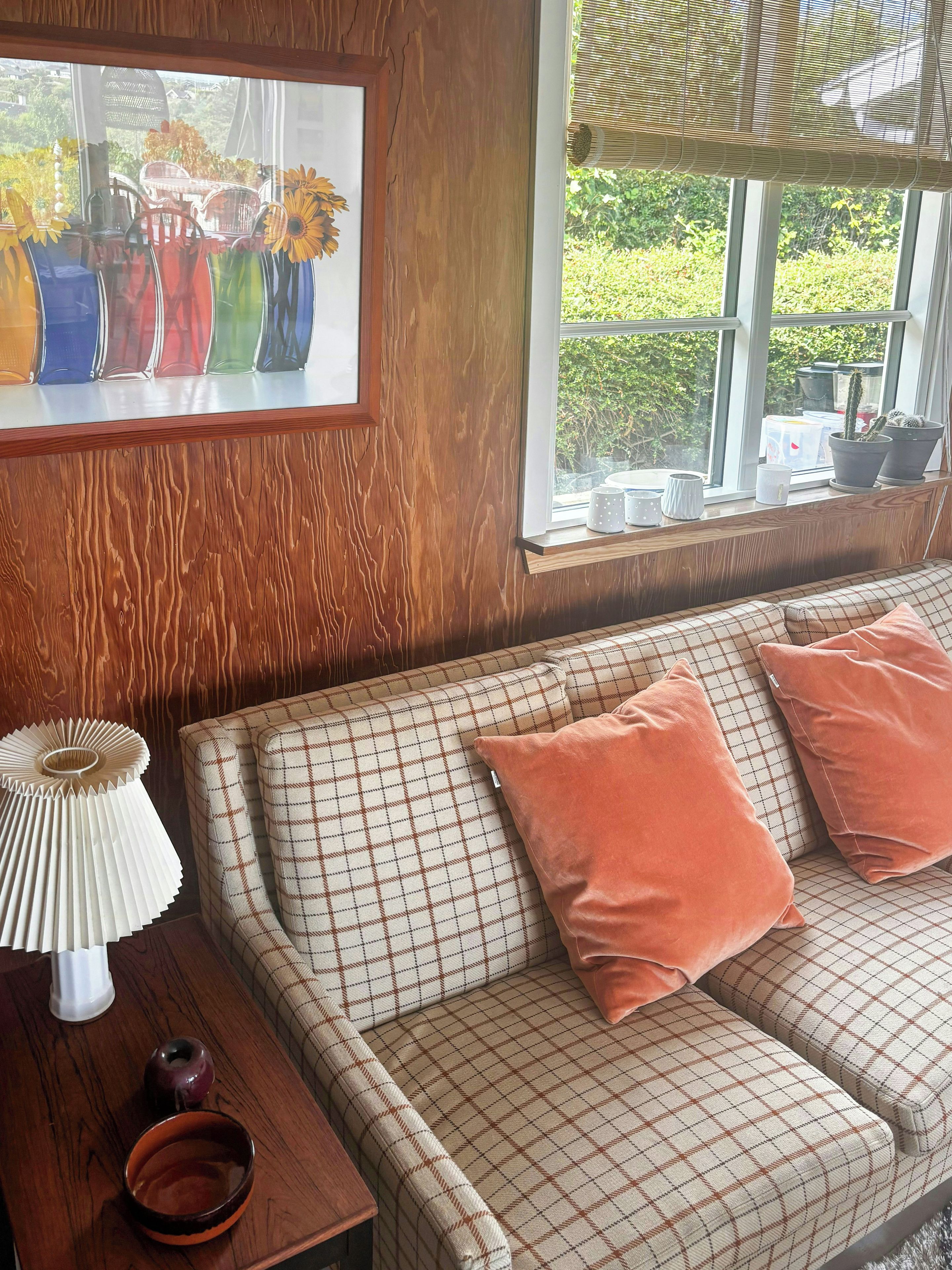 Una sala de estar acogedora con un sofá a cuadros, una mesita de madera con una lámpara y una ventana con vistas a zonas verdes.