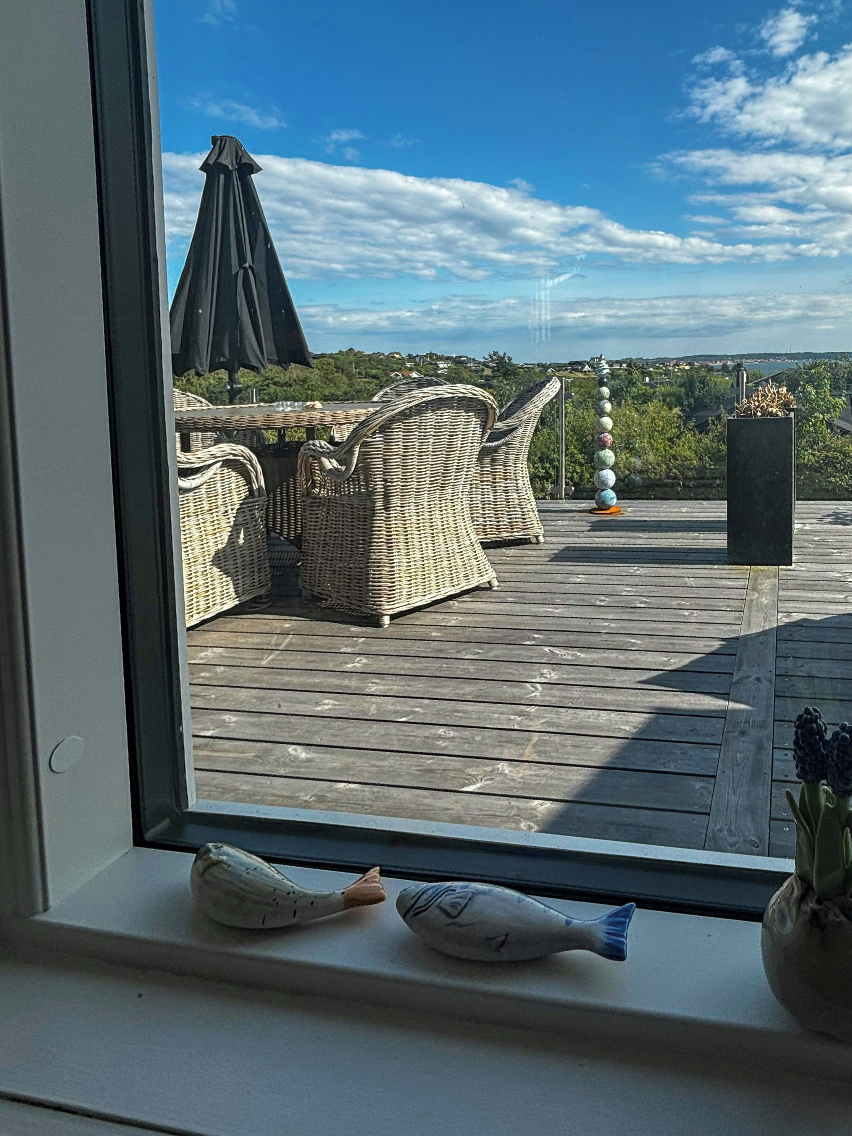 Une vue à travers une fenêtre donnant sur une terrasse en bois, avec des meubles en osier et une colonne décorative.