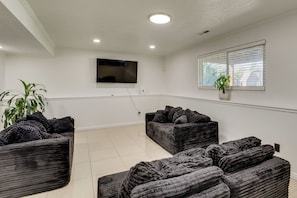 A room with a flat-screen TV mounted on the wall, a window with blinds, and several dark-colored sofas arranged in a semi-circle. - Gateway to Mountain Fun! 5BR Utah Home (Lehi)