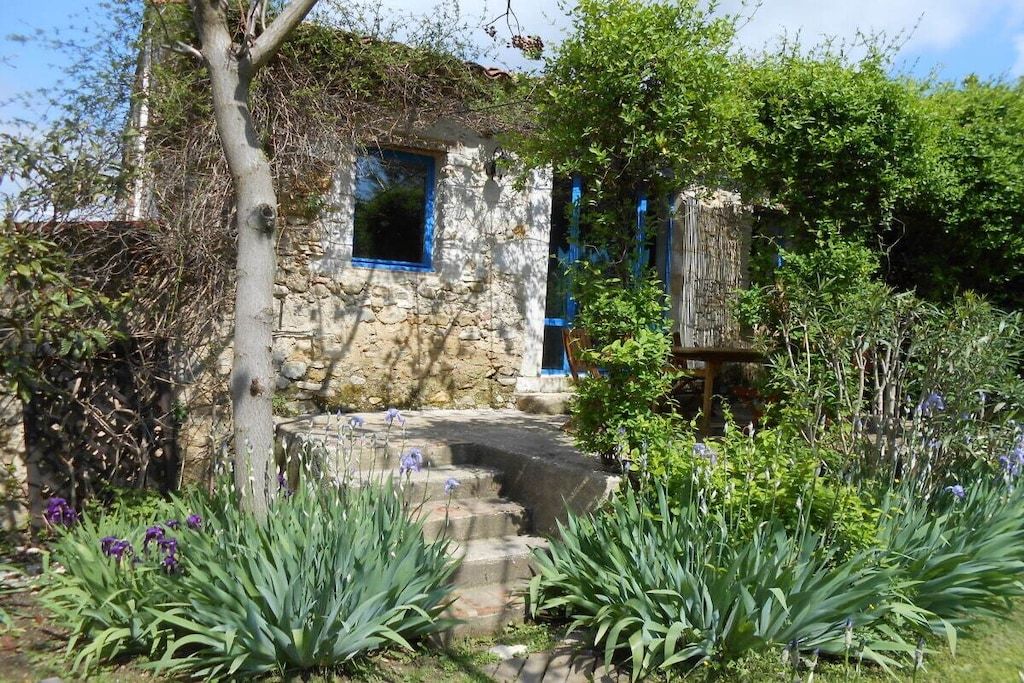 Edificio de piedra con ventana y puerta azules, rodeado de abundante vegetación y un jardín con diversas plantas.