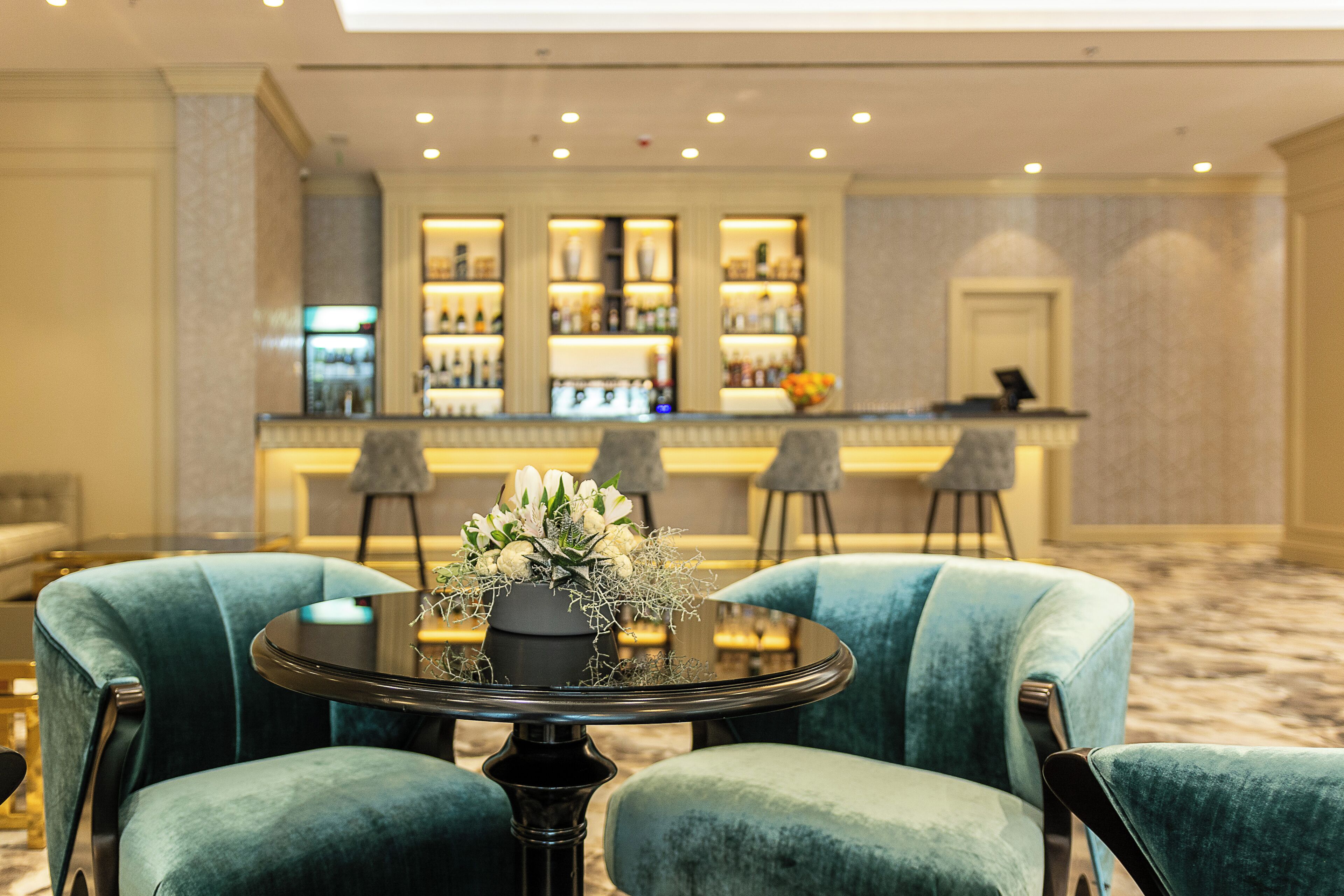 A bar area with a counter and shelves of bottles, two green armchairs, and a table with a floral centerpiece.