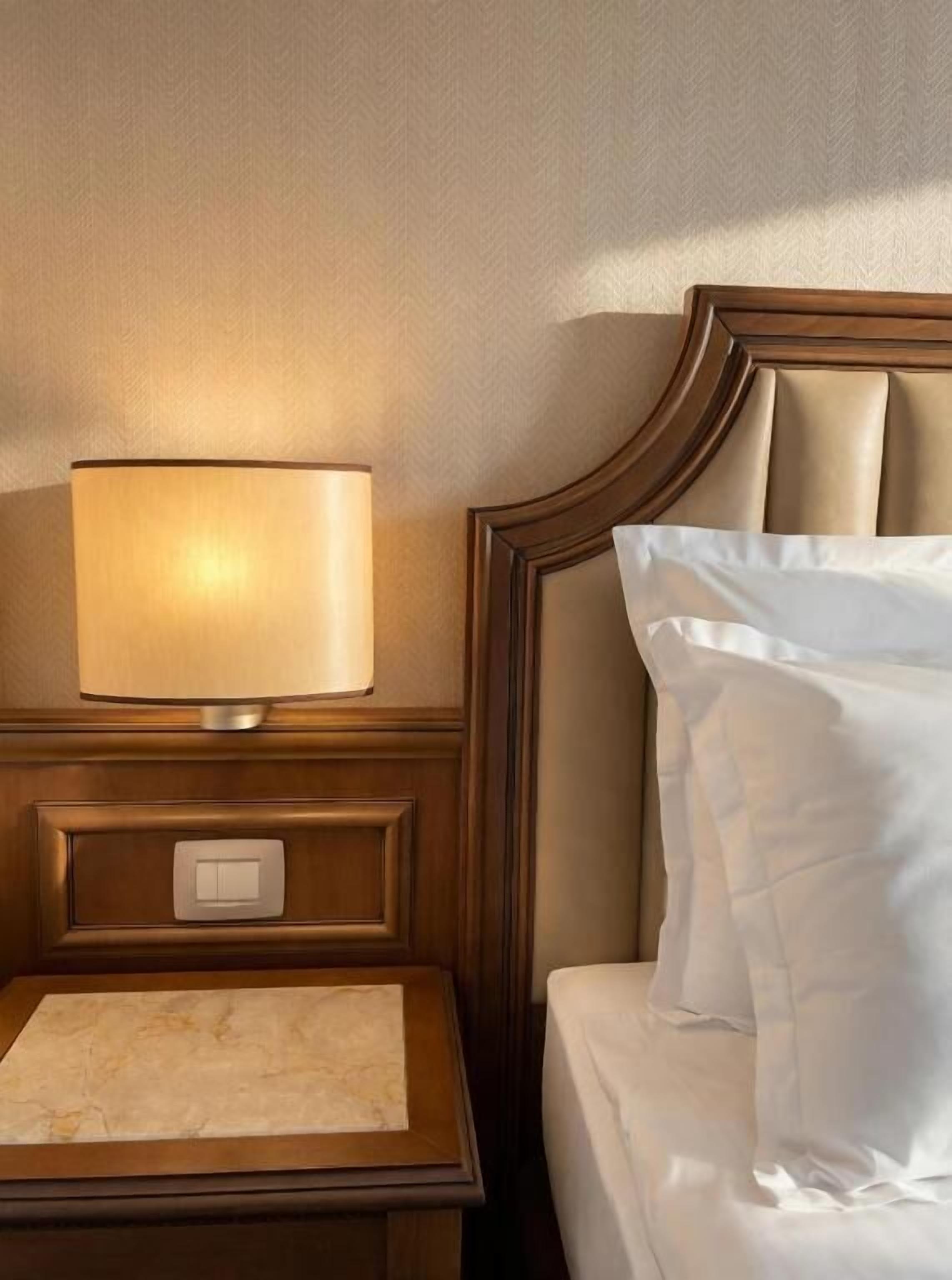 A hotel room with a wooden nightstand, a lamp, a bed with white linens, and a headboard.