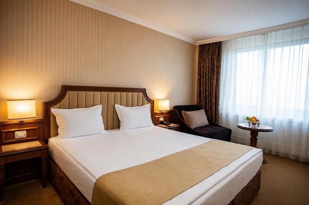 A hotel room with a large bed, two bedside tables with lamps, a chair, a small round table with fruit, and a window with curtains.