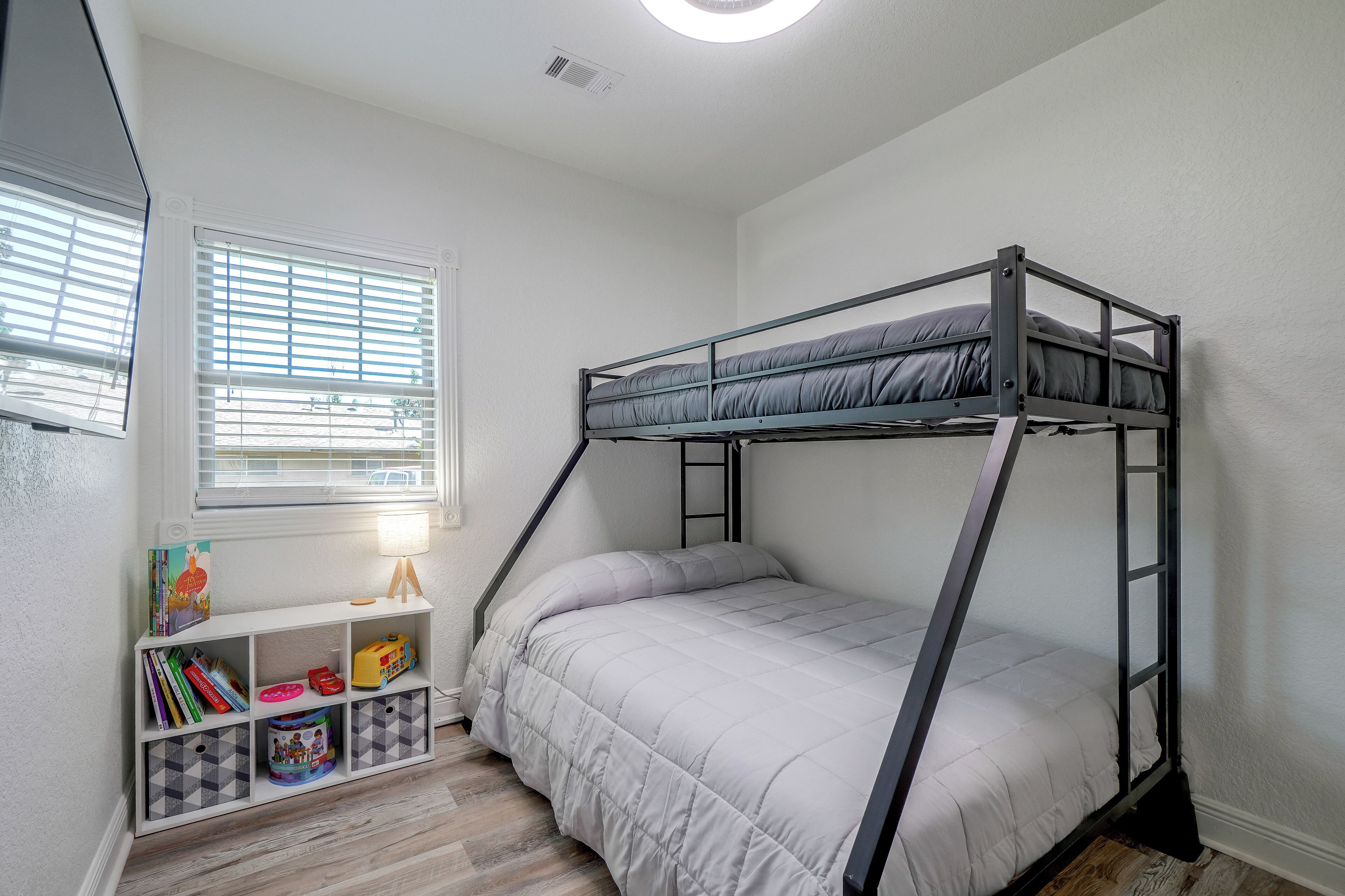 A bunk bed with a white mattress and grey bedding, a white shelving unit with books and toys, and a window with blinds.