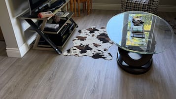 A living room with a flat-screen TV, a glass coffee table, a patterned armchair, a small wooden table with a lamp, and a cowhide rug.