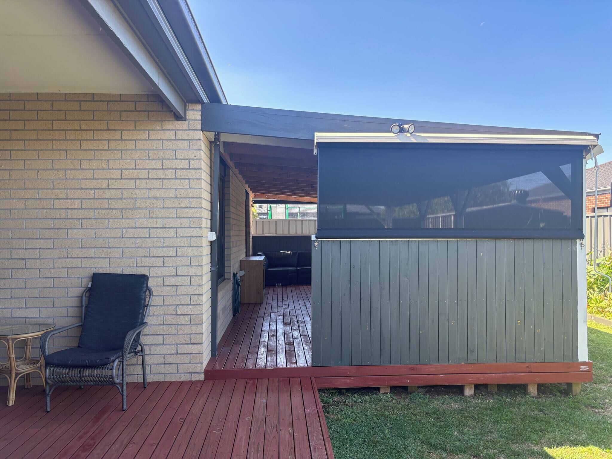 A covered patio area with a wooden deck, a seating area with a chair and table, and a fenced enclosure.