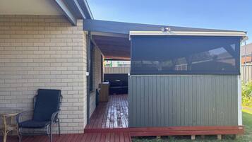 A covered patio area with a wooden deck, a seating area with a chair and table, and a fenced enclosure.