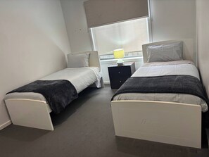 A small, single-room hotel bedroom with two single beds, a bedside table with a lamp, and a window with blinds.