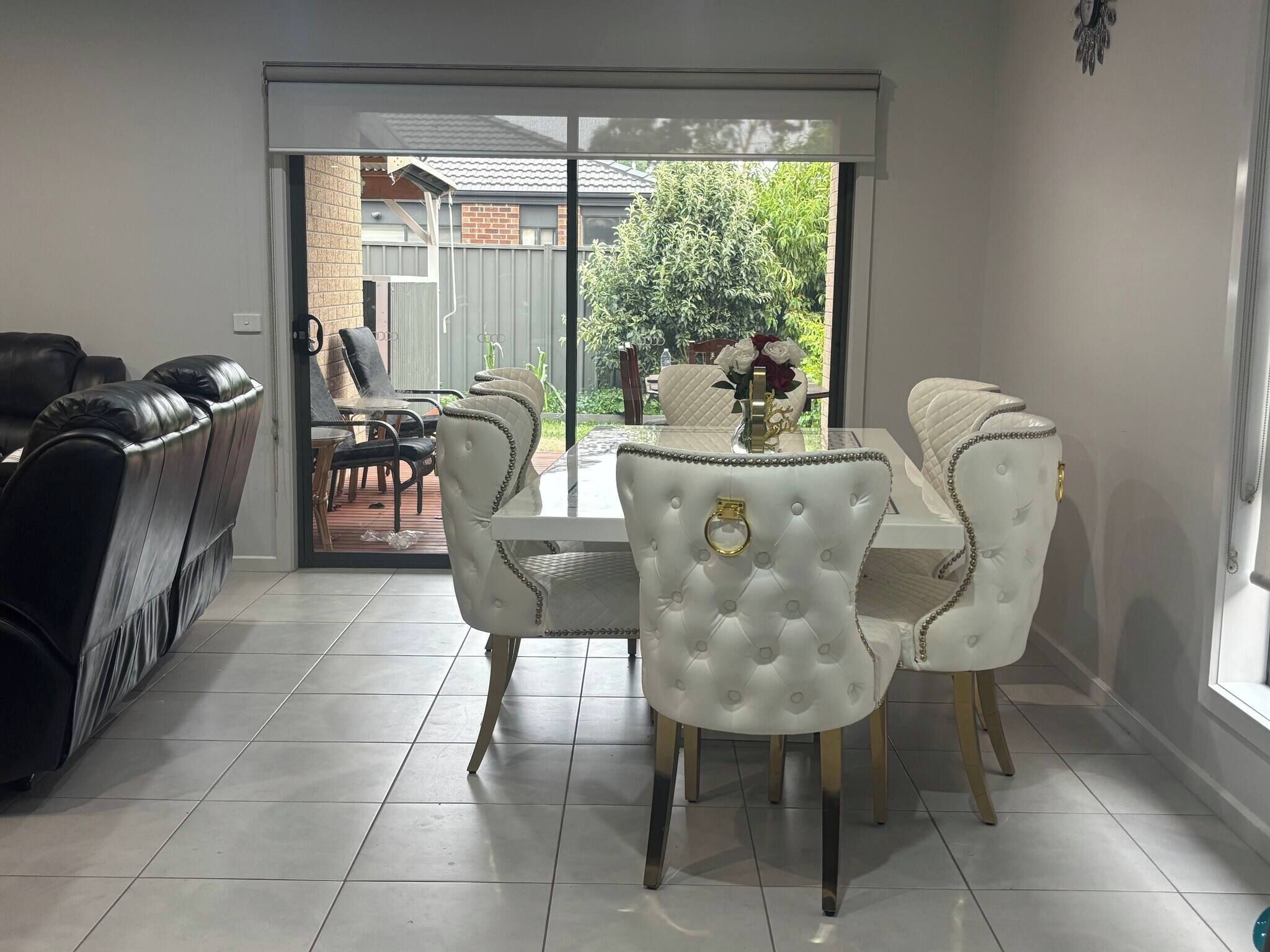 Un coin repas avec une table et des chaises blanches, un canapé noir et une vue sur l’extérieur par une porte vitrée.