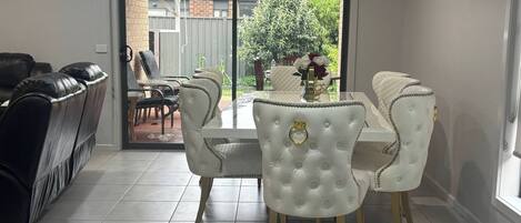 Un coin repas avec une table et des chaises blanches, un canapé noir et une vue sur l’extérieur par une porte vitrée.