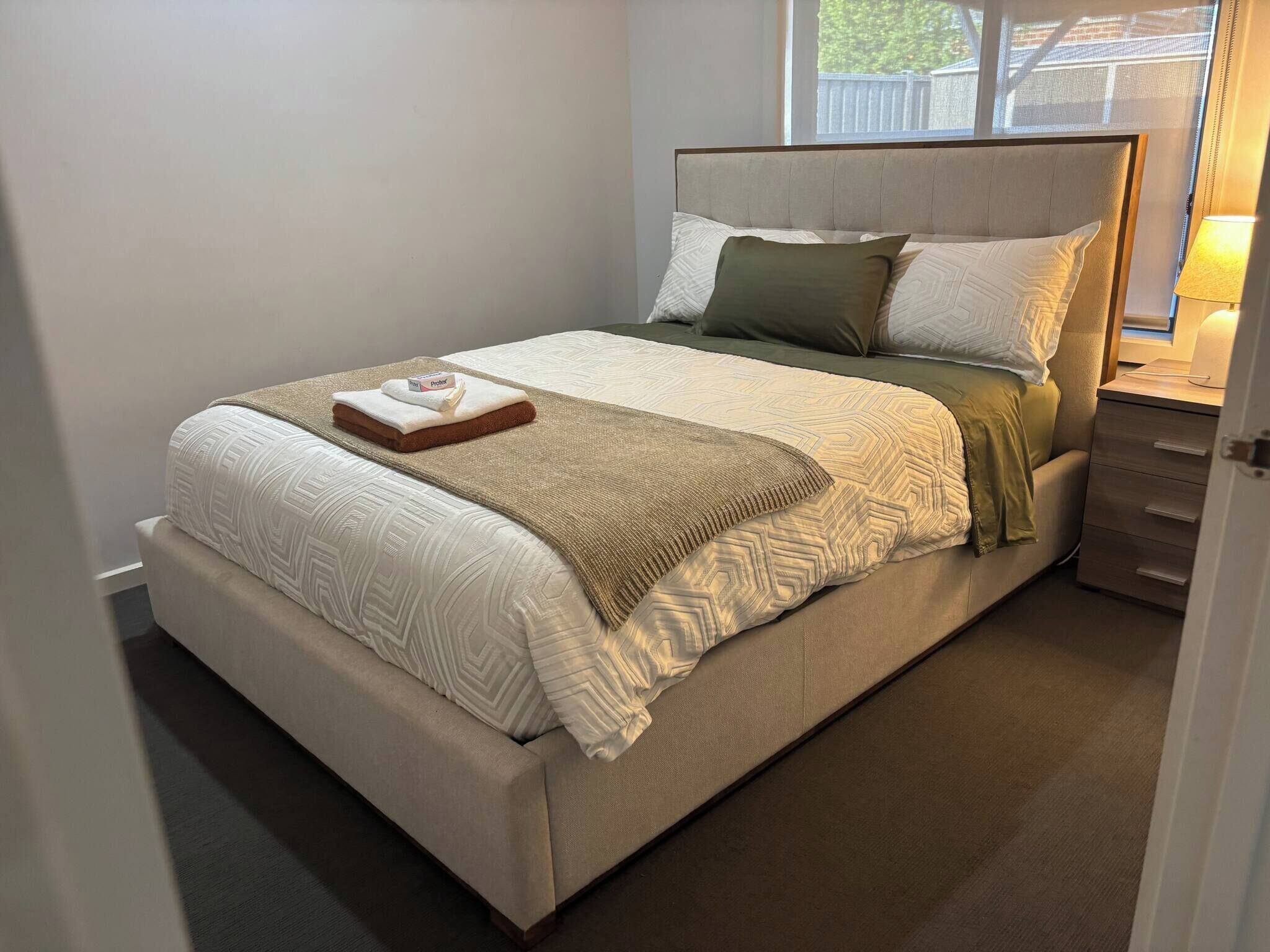 A neatly made bed with a geometric patterned bedspread, a folded towel, and a box of tissues.