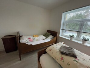 Ein Einzelbett mit Blumenmuster auf der Bettwäsche, ein Nachttisch, ein Fenster mit Jalousien und ein kleiner Topfpflanzen auf dem Fensterbrett.