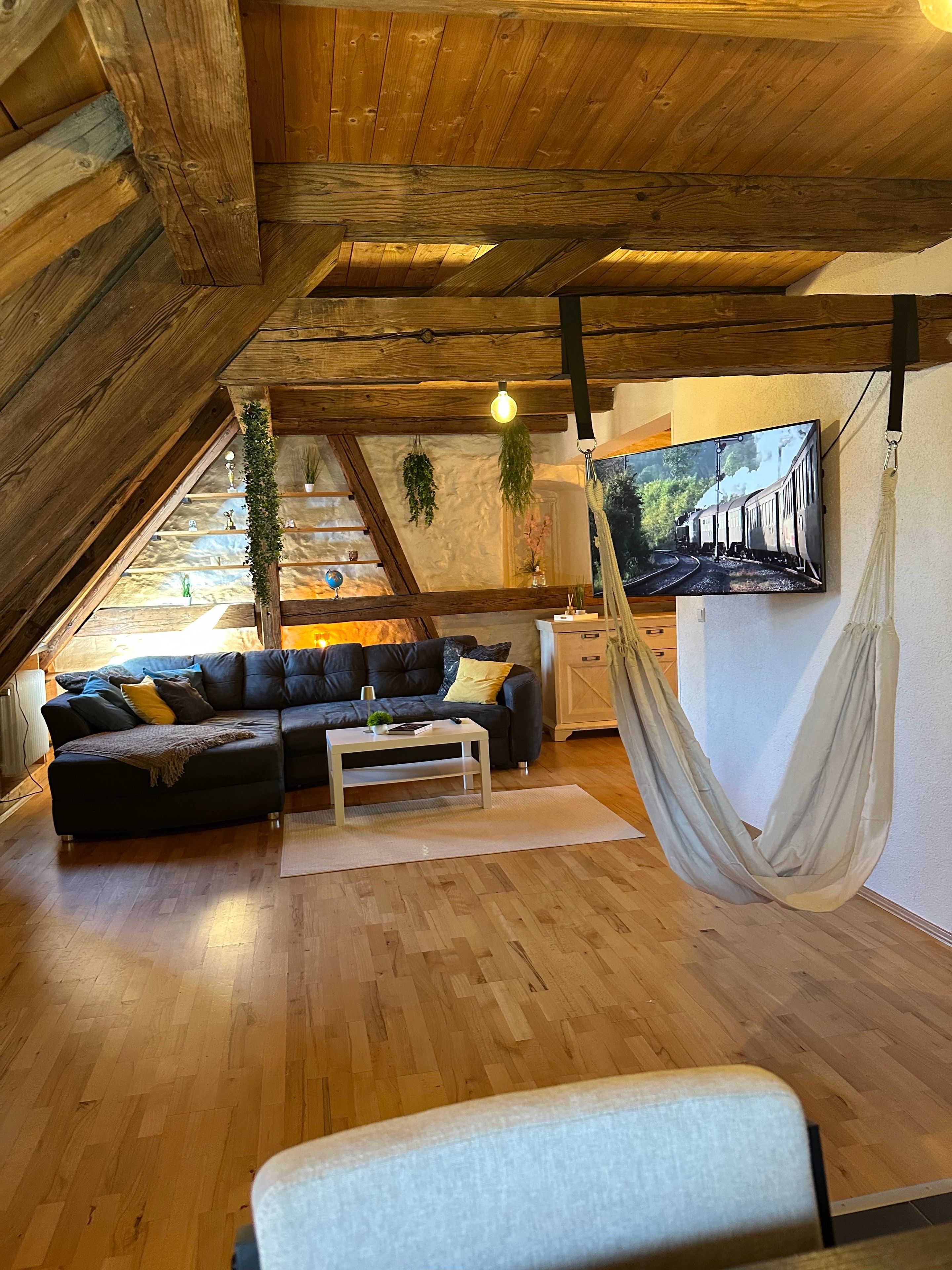 A cozy loft-style living room with a wooden ceiling, a black sectional sofa, a white coffee table, and a hammock.
