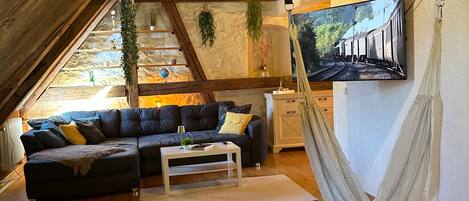 A cozy loft-style living room with a wooden ceiling, a black sectional sofa, a white coffee table, and a hammock.