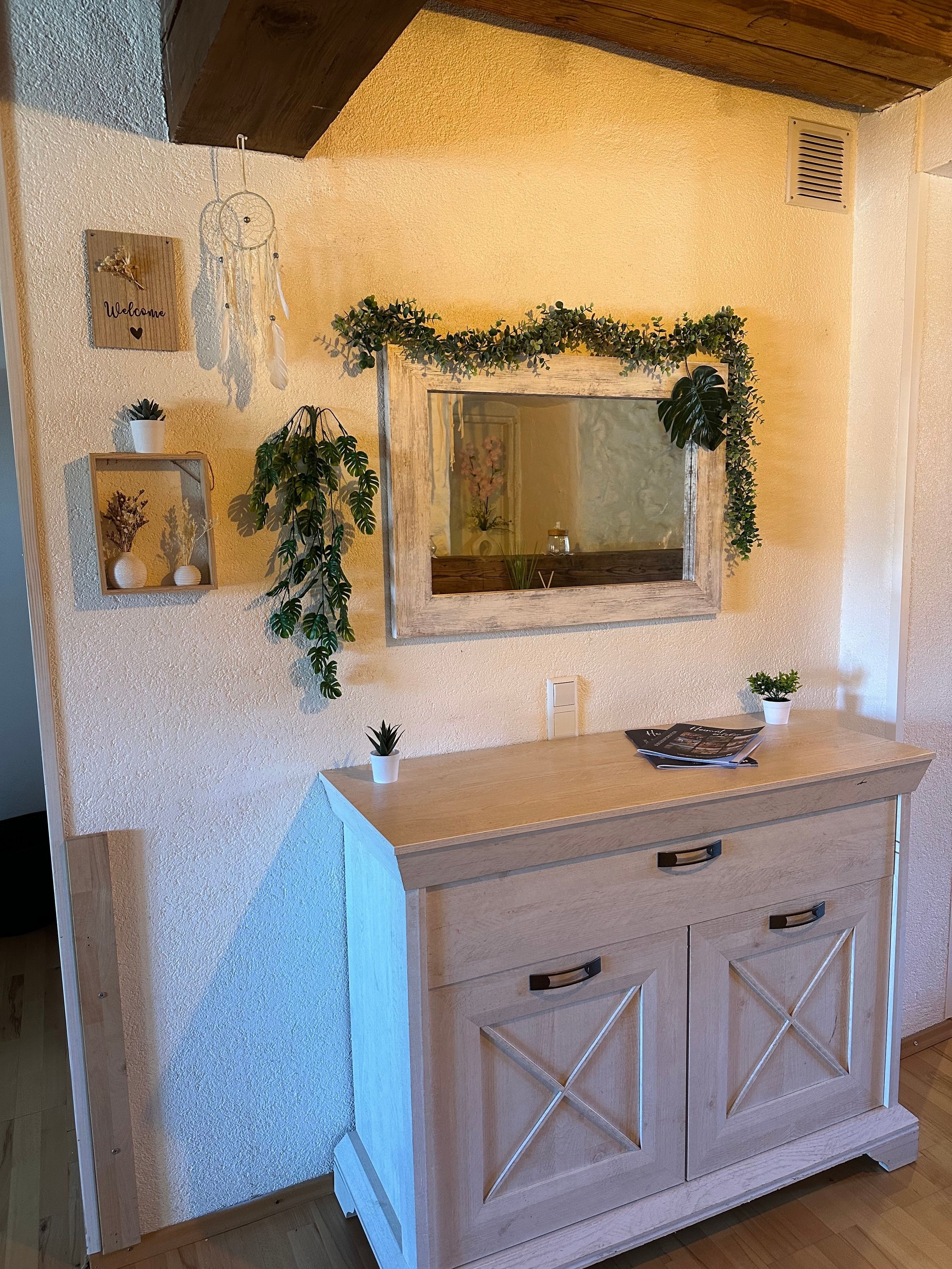 A wooden vanity with a mirror, a small plant, and a welcome sign on the wall.