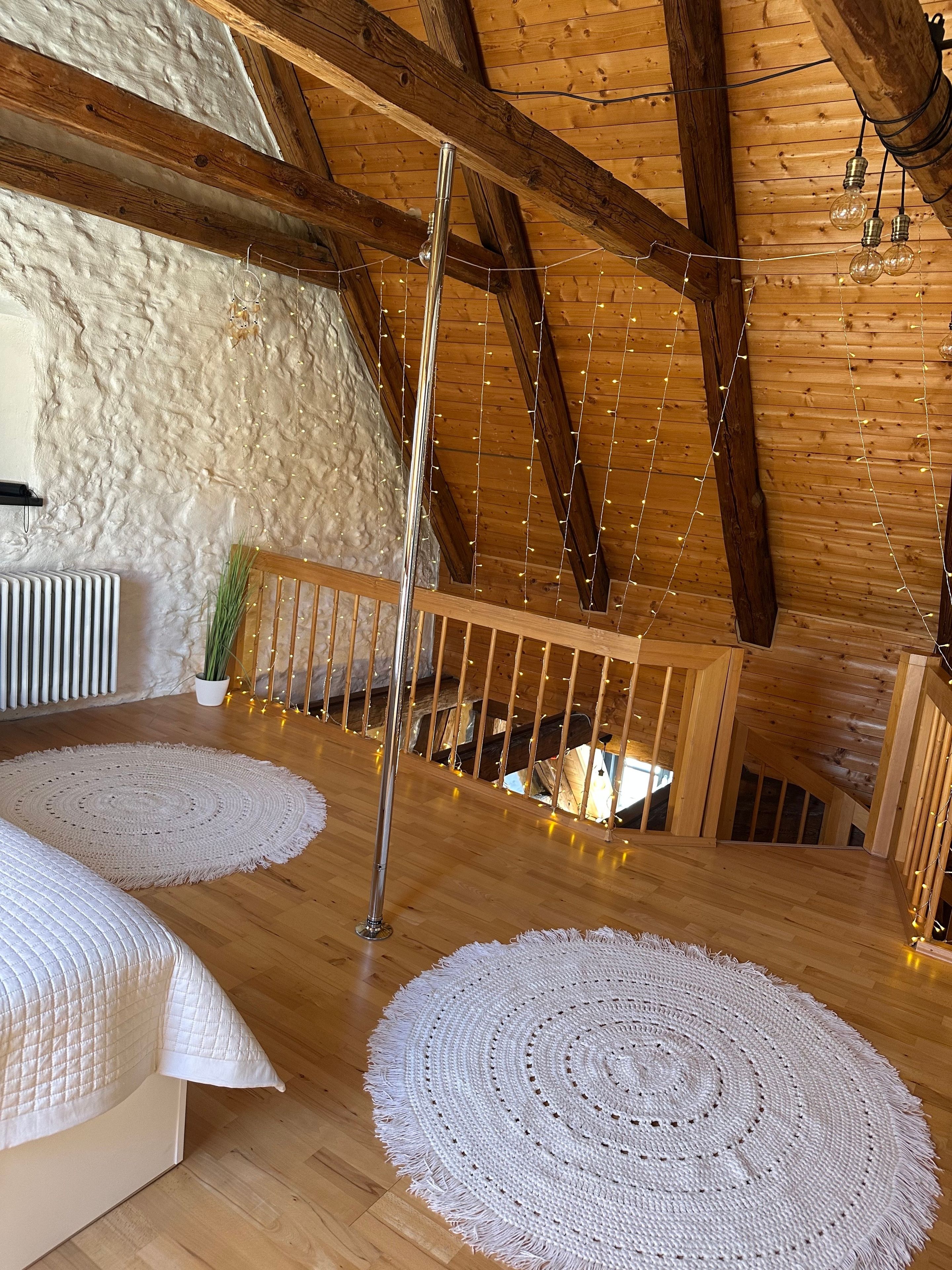 Ein Zimmer mit Holzbalken, hoher Decke, einem Bett mit weißer gesteppter Bettdecke, zwei Häkelteppichen und Lichterketten.