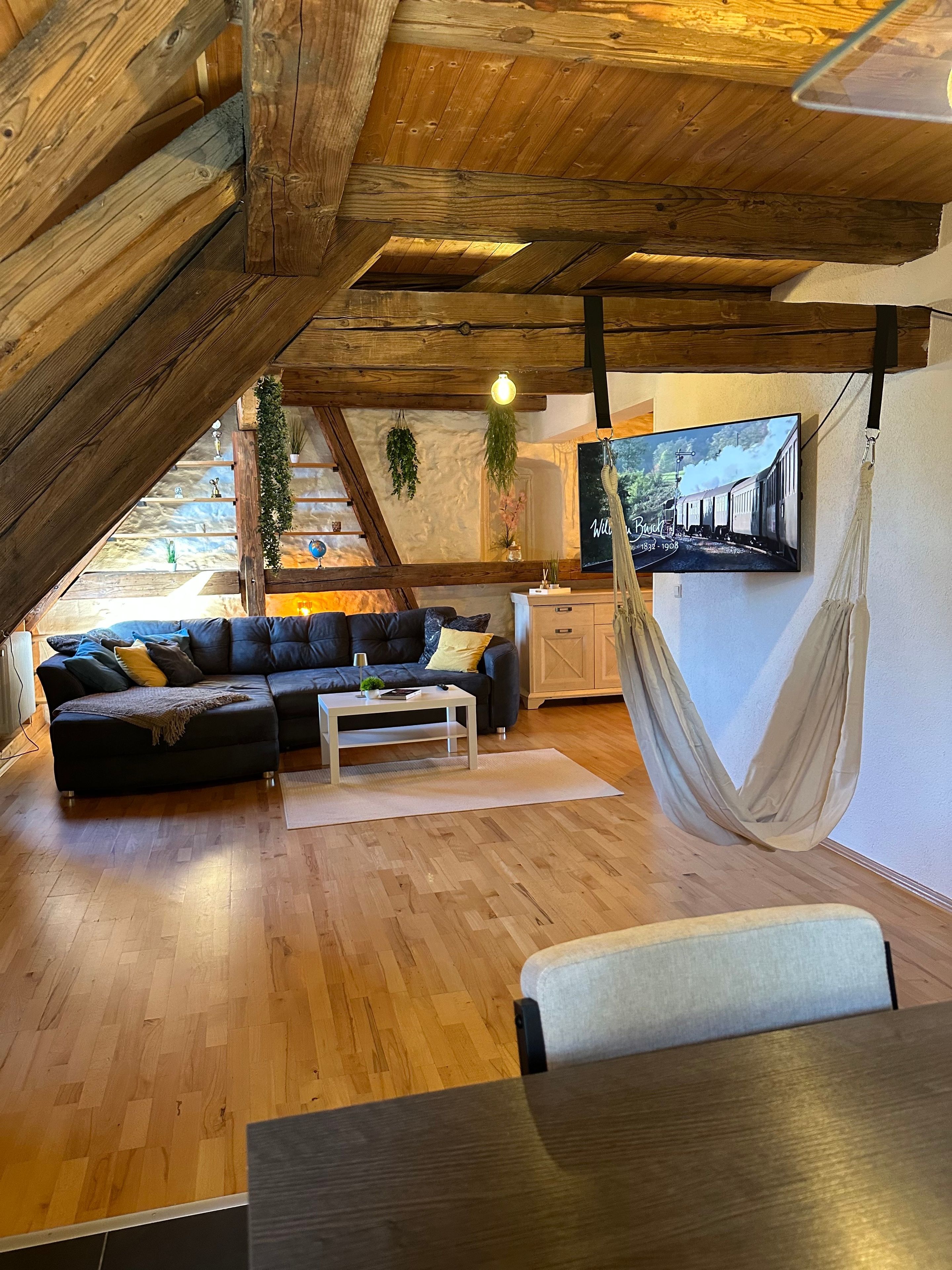 Ein Loft-Wohnzimmer mit Holzbalkendecke, einem schwarzen Ecksofa, einem weißen Couchtisch und einer Hängematte.