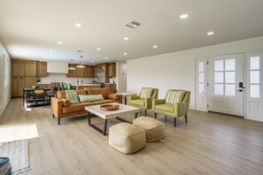 Ein geräumiger Wohnbereich mit einer Couch, Hocker und Sesseln, im Hintergrund eine Küche und eine Tür mit satinierten Glasscheiben.