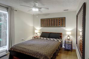 Ein Schlafzimmer mit einem Bett, zwei Nachttischen mit Lampen, einem Deckenventilator, einem großen Fenster und einem Wandbild.