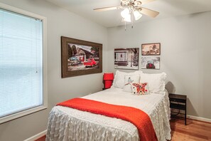 Une chambre à coucher comprenant un lit, un ventilateur de plafond, une fenêtre avec des stores et des tableaux accrochés au mur.