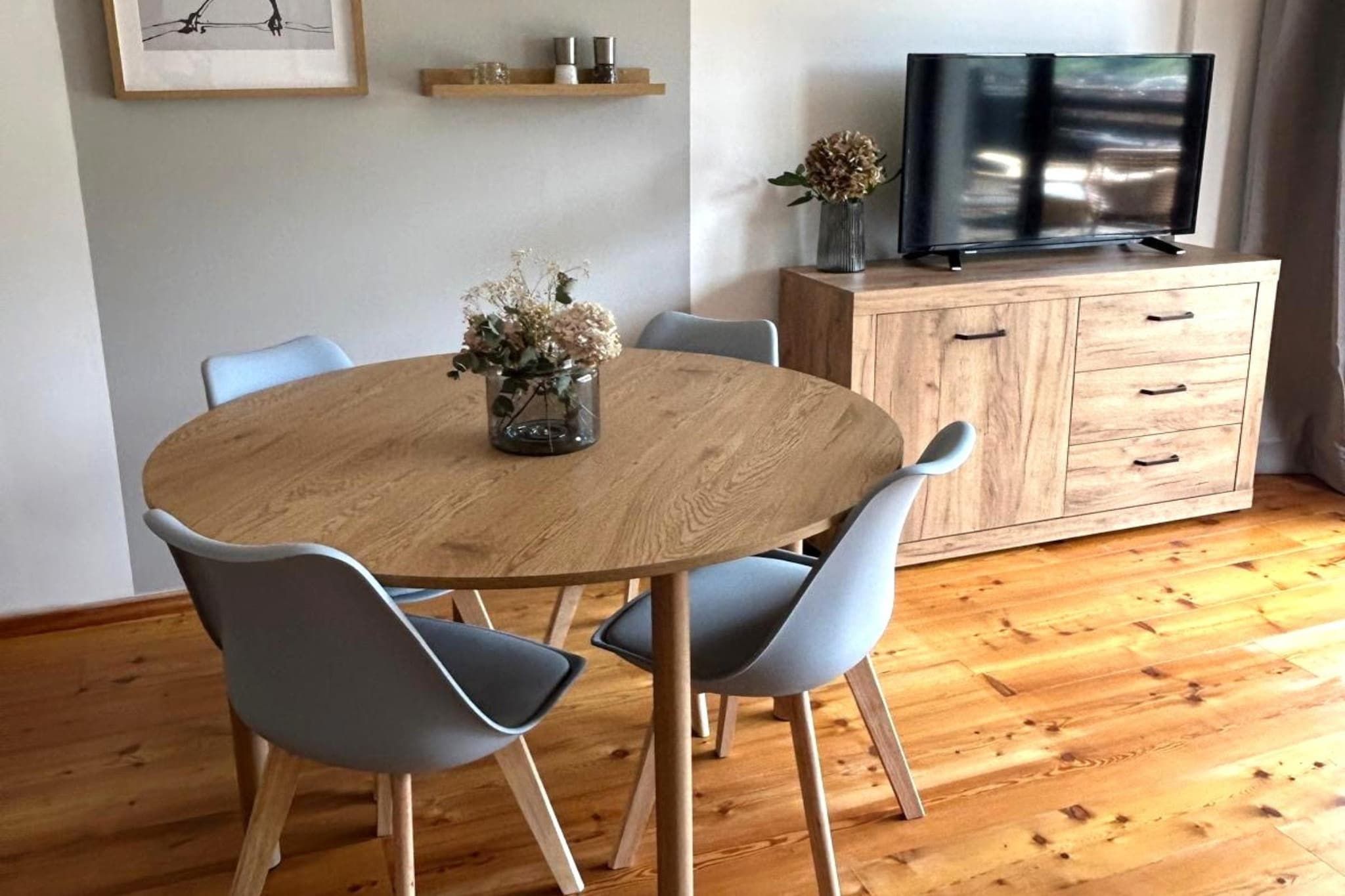 Een ronde houten eettafel met vier stoelen, een vaas met bloemen, een houten kast met laden en een televisie erbovenop.