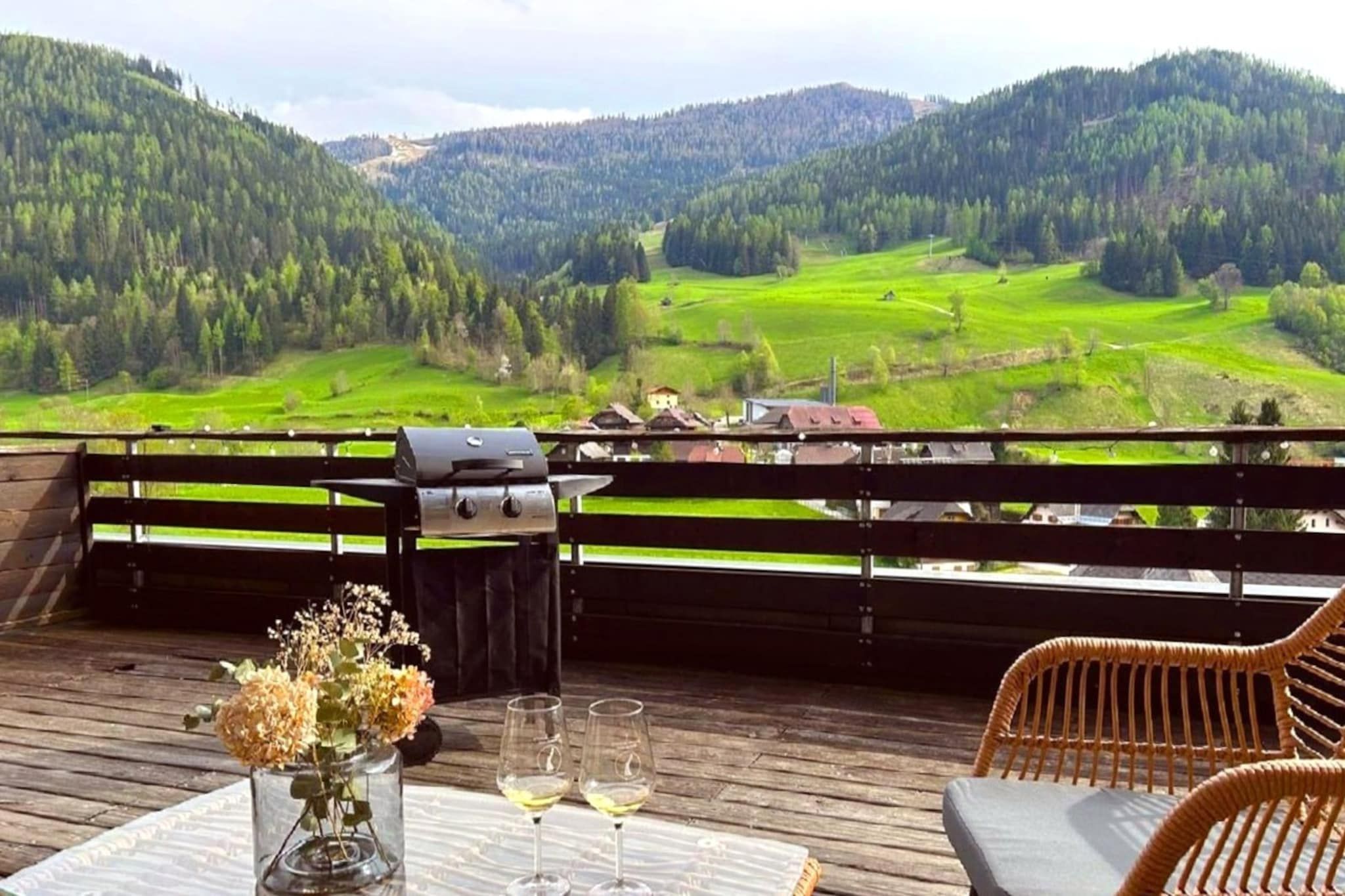Un terrazzo in legno con una griglia per barbecue, un tavolo con un vaso di fiori e due bicchieri da vino.