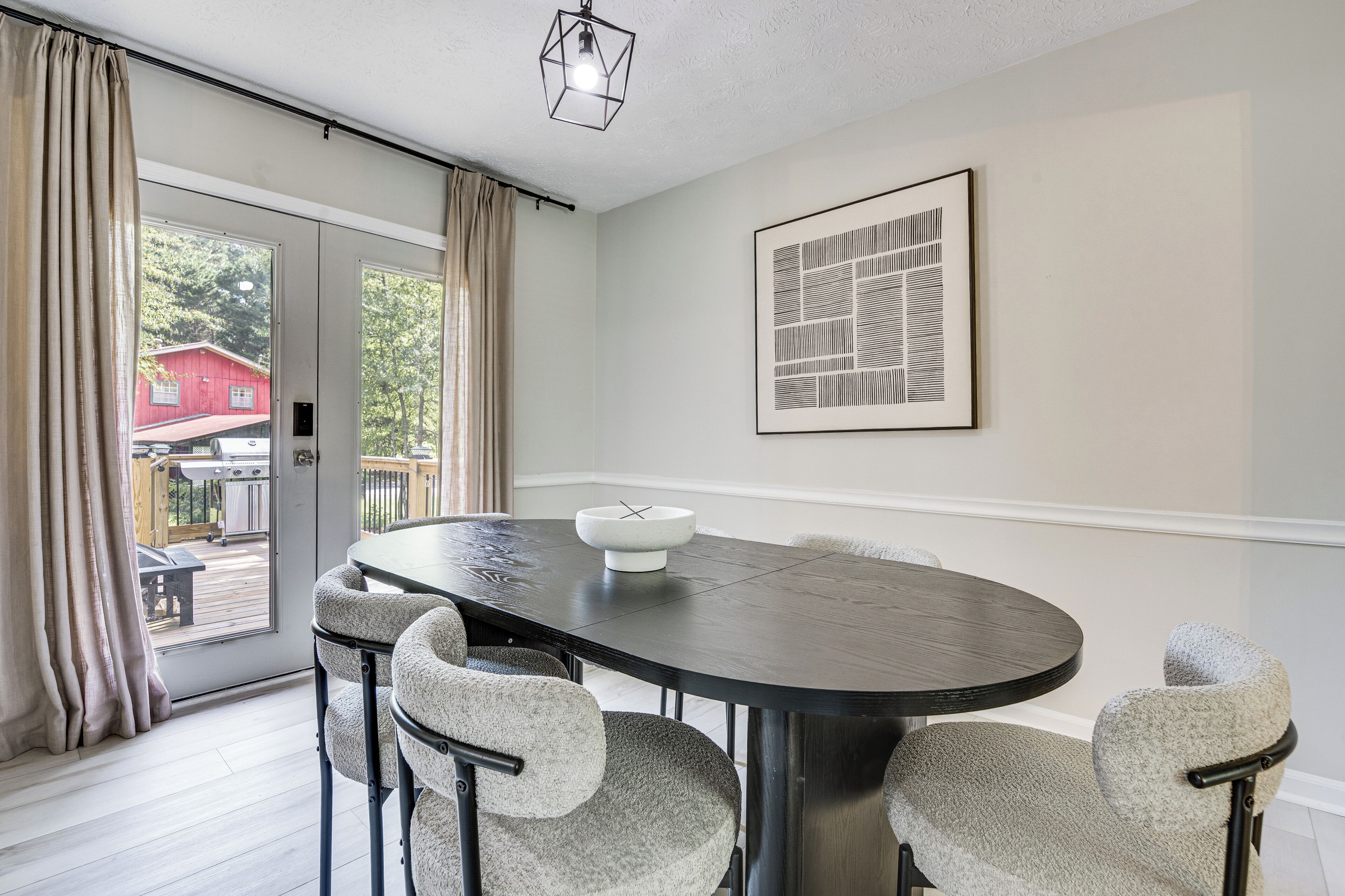 Un coin repas comprenant une table ronde, quatre chaises, un luminaire, une porte avec des rideaux et une œuvre d’art abstraite encadrée accrochée au mur.
