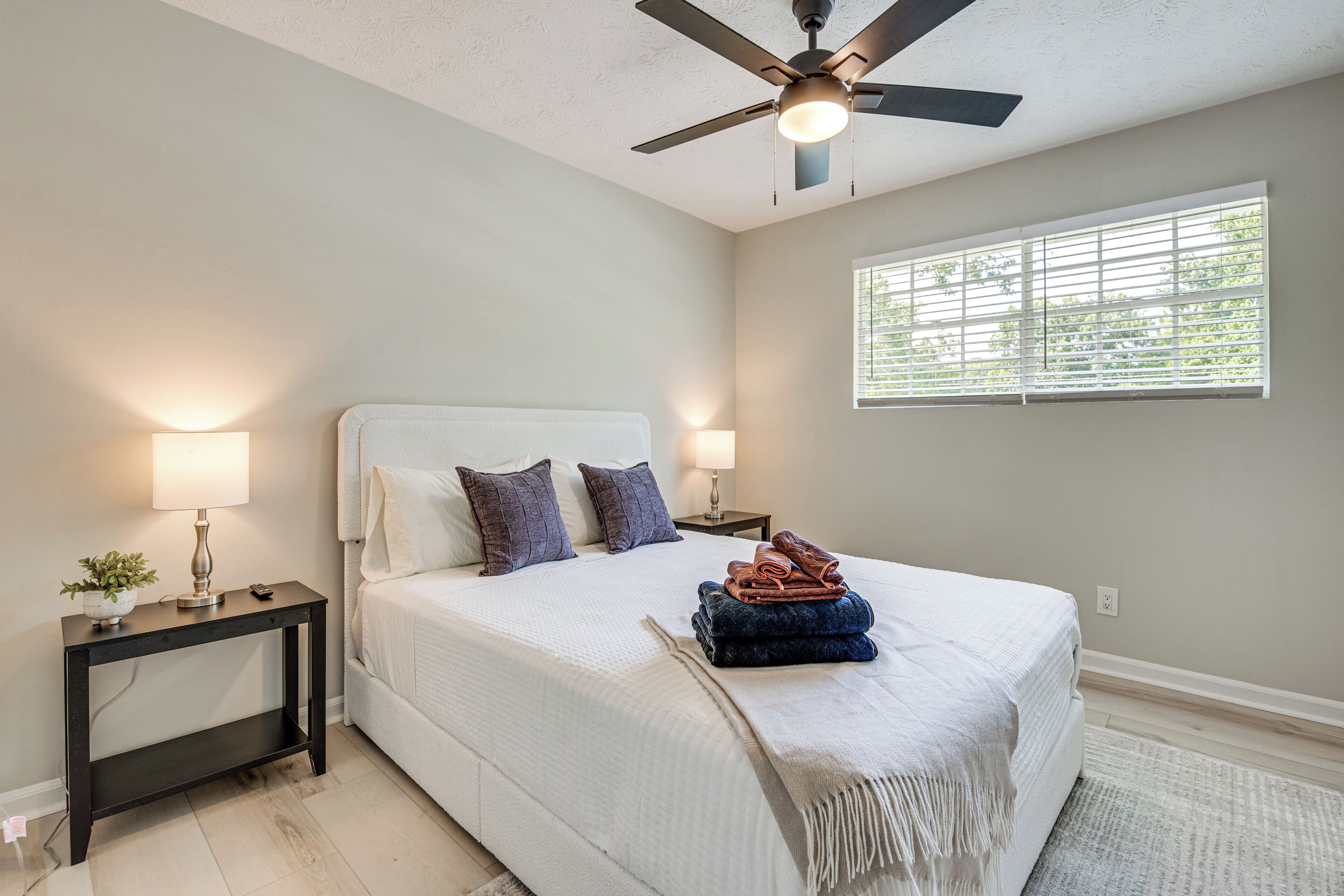 A bedroom with a bed, a nightstand, a lamp, a ceiling fan, and a window with blinds.