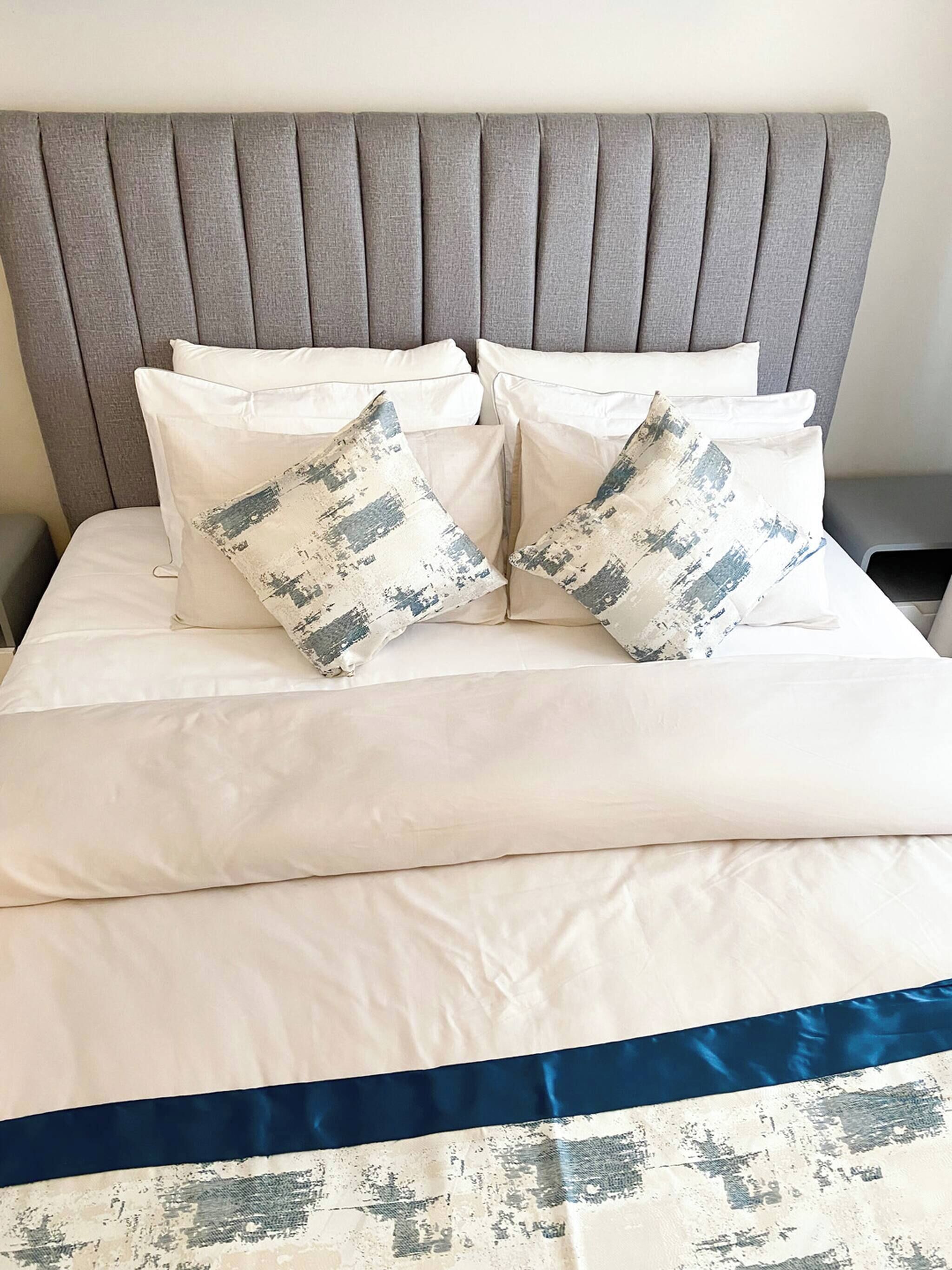 A bed with a grey upholstered headboard, white bedding, and two decorative pillows with a distressed design.
