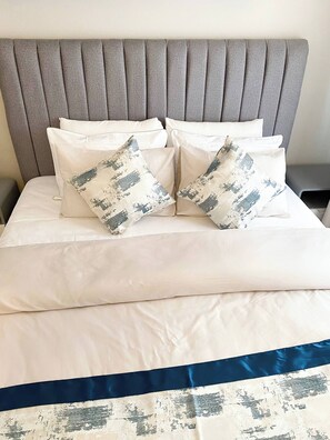 A bed with a grey upholstered headboard, white bedding, and two decorative pillows with a distressed design.
