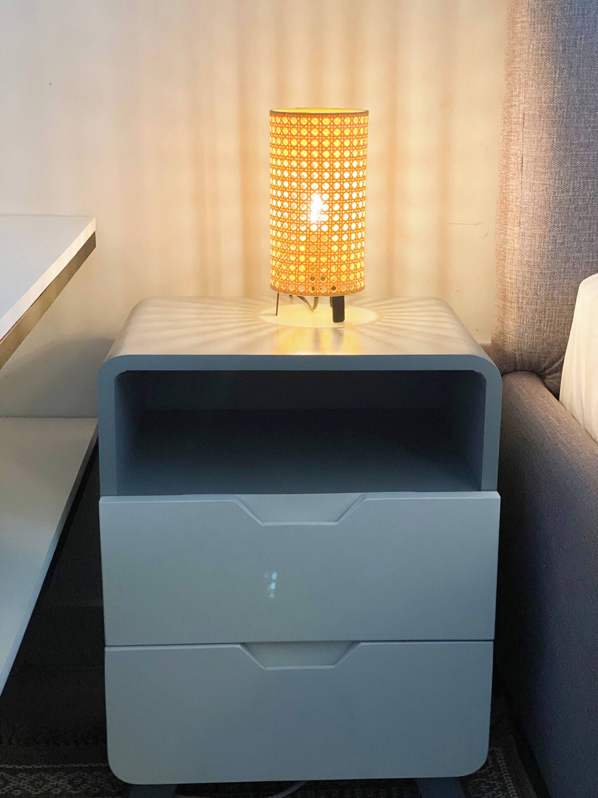 Un comodino moderno con una lampada, un cassetto e un divano grigio sullo sfondo.