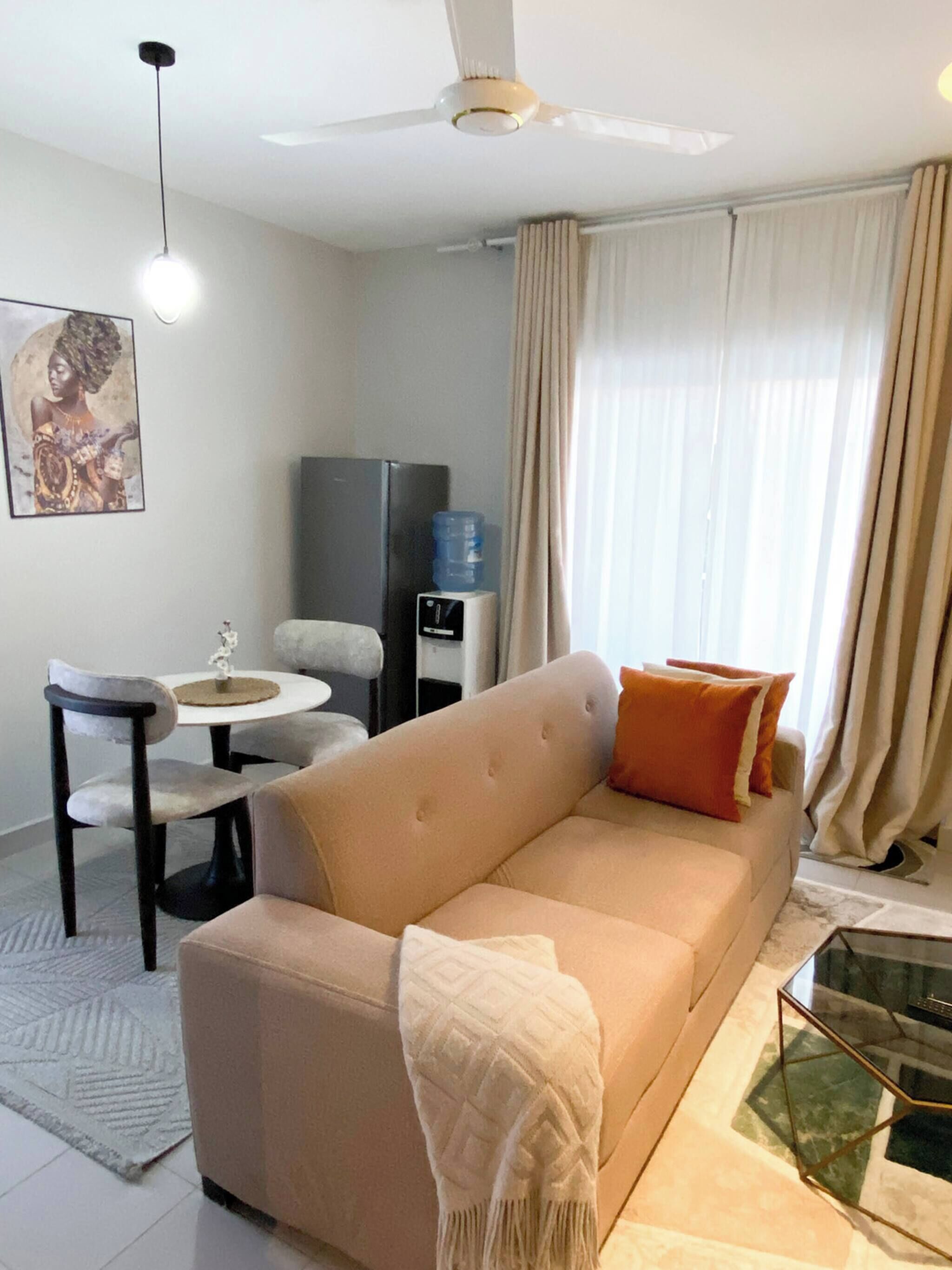 Un soggiorno con un divano beige, un tavolino in vetro, un'area pranzo con tavolo e sedie, un distributore d'acqua e una ventola a soffitto.