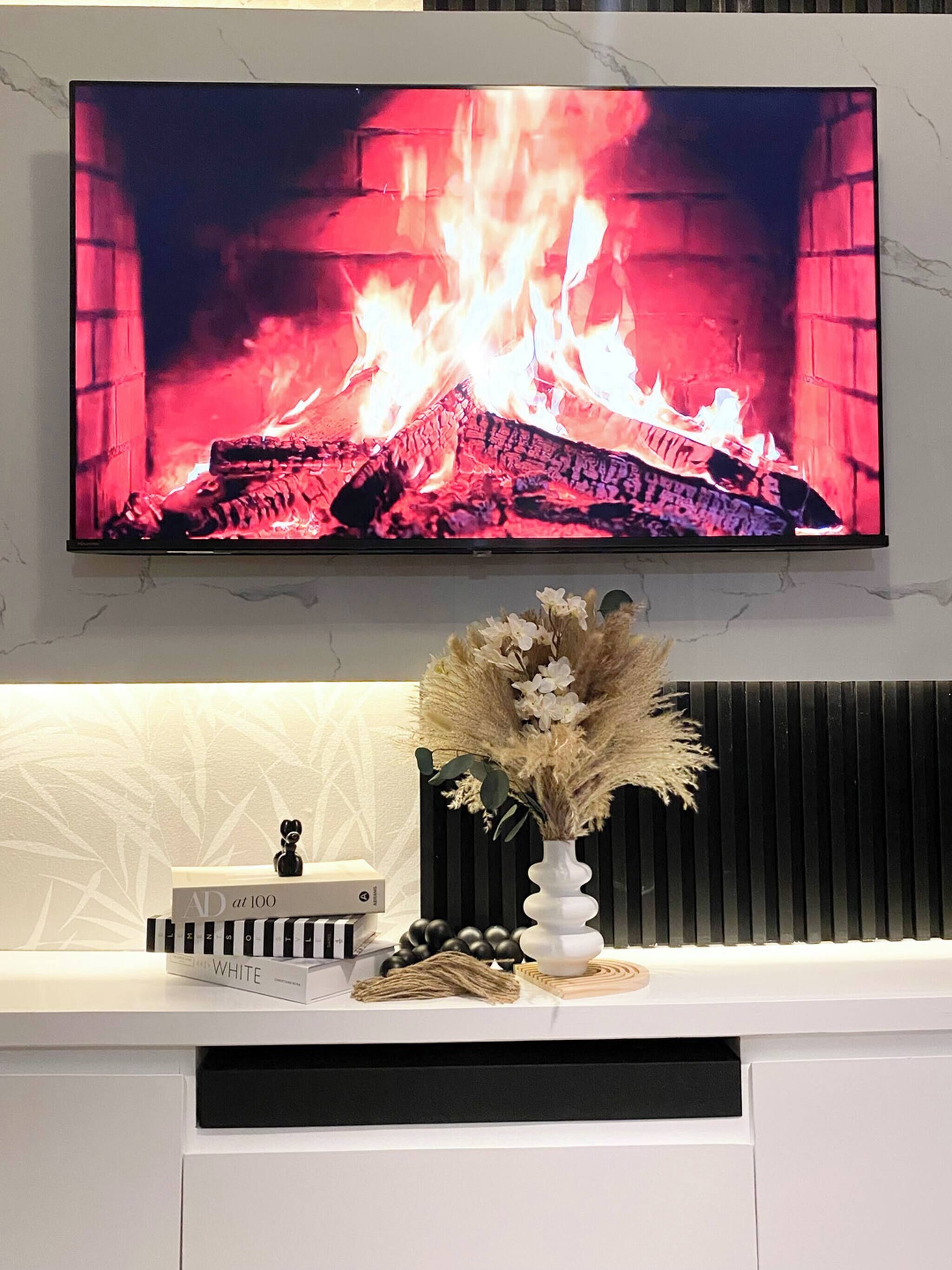 A flat-screen TV mounted on a wall above a white console with a decorative vase and a book.