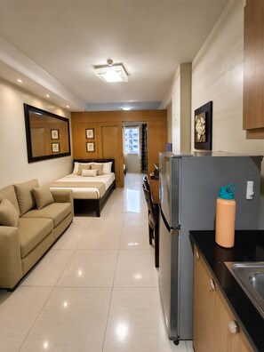 A modern hotel room with a bed, a sofa, a kitchen area with a refrigerator, and a bathroom visible through an open door. - Modern 2-Bedroom Apartment with Shared Pool in Central Pasay (Pasay City)