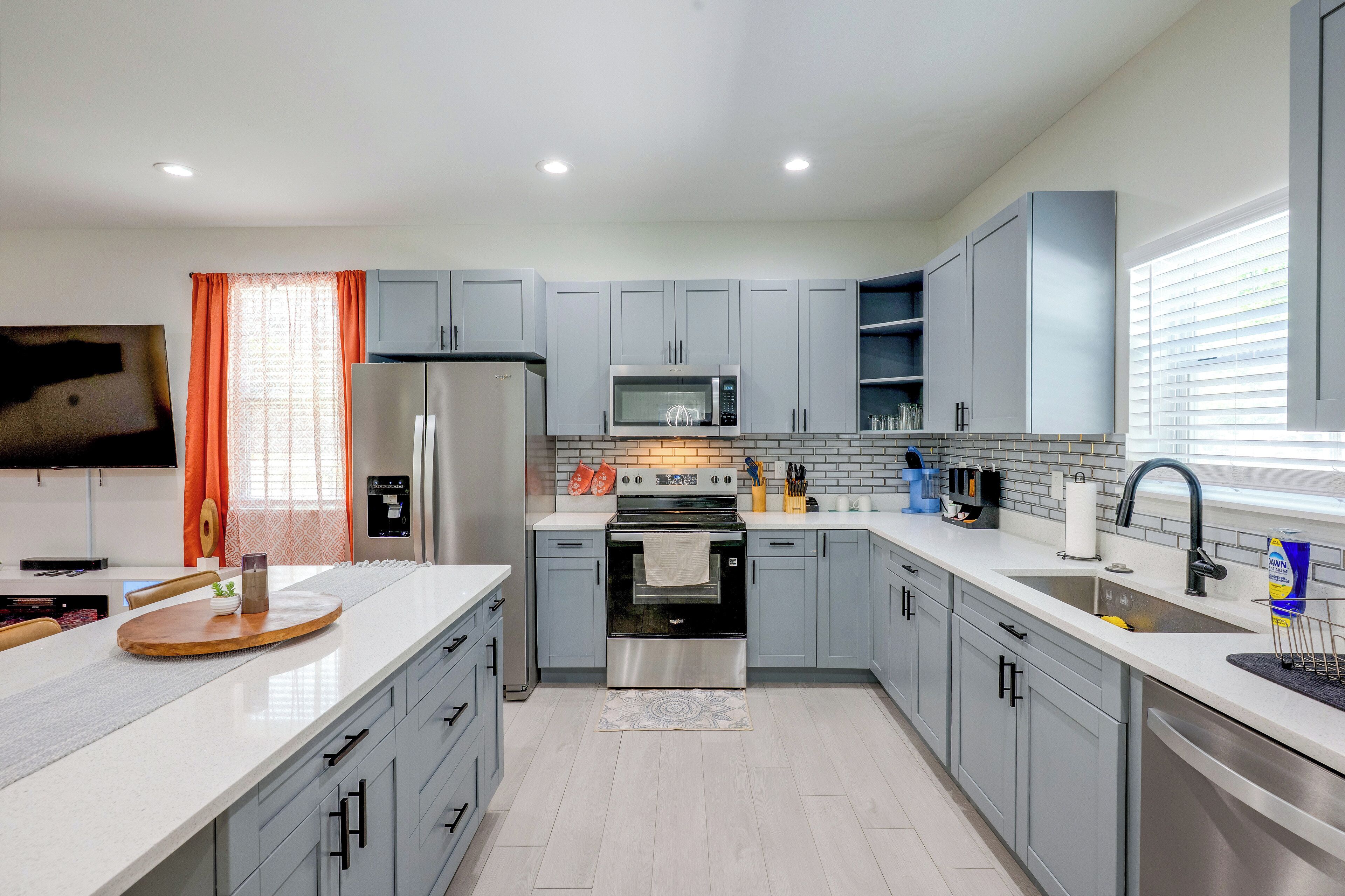 A modern kitchen with grey cabinets, stainless steel appliances, and a flat-screen TV mounted on the wall.