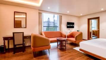 Habitación de hotel con cama, dos sofás, una mesita, un escritorio, una silla, televisor y ventana con vistas a edificios y árboles.