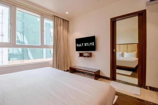 Habitación de hotel con cama, televisión instalada en la pared, espejo y ventana con cortinas.