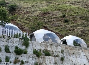 Tres estructuras de cúpulas geodésicas en una colina, con una cerca blanca de piquetes al frente.